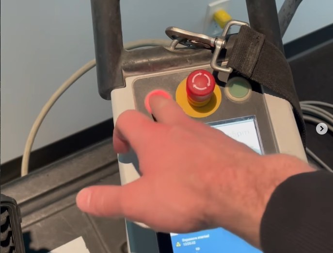 An operator's hand on the red emergency-stop button of the digital X-Ray control panel — cutting power to the source stops radiation instantly