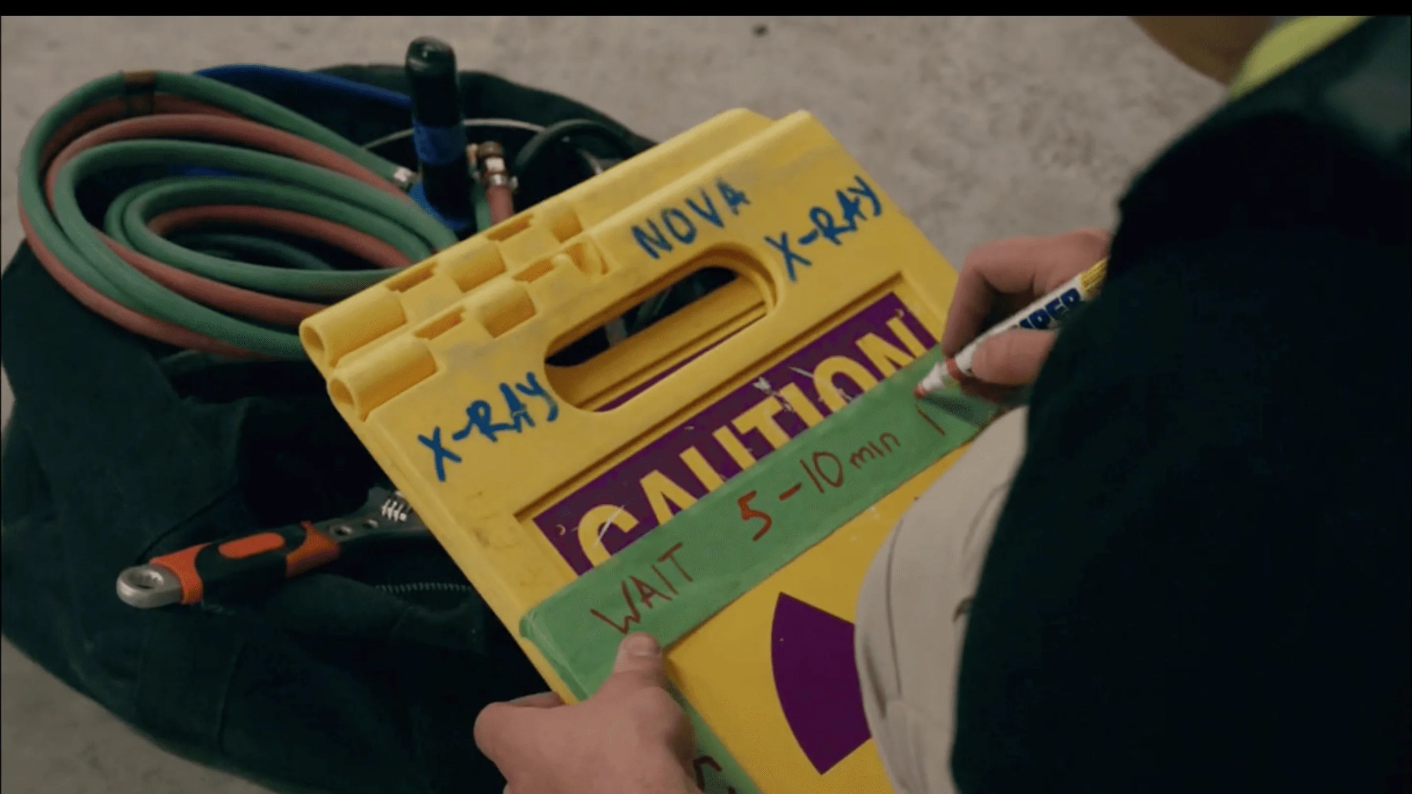 A crew member hand-labeling a yellow Nova X-Ray caution barrier with a marker — 'WAIT 5–10 min' — next to coiled hoses and tools staged on site